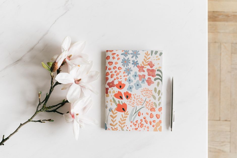 A floral notebook, pen and pale flowers arranged on a light desk for a quiet reading and note-taking routine.